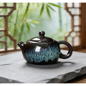 May include: A small, dark brown ceramic teapot with a glossy finish and a teal and blue glaze. The teapot has a rounded body, a curved handle, and a small spout. It sits on a gray stone surface, with a blurred background of bamboo and a wooden window.