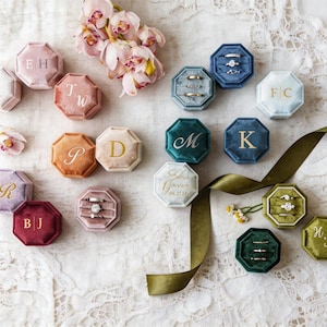 Op de afbeelding: Een assortiment fluwelen ringendoosjes in diverse kleuren, waaronder roze, blauw en groen, gerangschikt op een wit kanten oppervlak. Elk achthoekig doosje heeft een gouden initiaal of inscriptie. Sommige doosjes bevatten ringen.