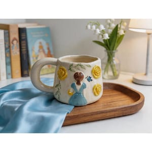 Peut inclure: Mug en céramique crème avec une grande anse, décoré de roses jaunes, de feuilles vertes et d'une figurine en robe bleue. Un papillon bleu est également présent. Le mug est posé sur un plateau en bois, suggérant un article décoratif ou cadeau.