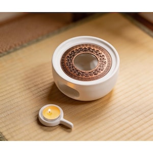 May include: A white ceramic tea warmer with a copper-colored decorative top and a small handle. A lit tea light candle sits next to the warmer. The tea warmer is designed to keep tea warm and is placed on a woven mat.