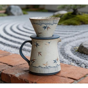 Puede incluir: Un juego de cafetera de cerámica para verter. La jarra y el portafiltro de color blanquecino están adornados con diseños de hojas azules y doradas. El juego incluye un cono de filtro a juego y se encuentra sobre una superficie de ladrillo rojo.