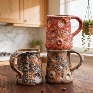 Può includere: Tre tazze in ceramica fatte a mano con superfici strutturate e design unici. Le tazze presentano una varietà di colori, tra cui rosso, arancione e blu. Ogni tazza ha un manico e un motivo distinto, simile a un cratere, che aggiunge fascino rustico.