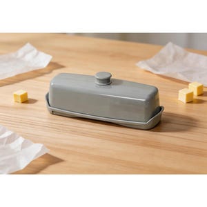 May include: A gray rectangular butter dish with a lid and a small knob on top. The dish sits on a matching tray. Several small cubes of butter and pieces of parchment paper are scattered around the dish on a wooden surface.