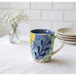 May include: A ceramic mug with a blue interior and a white handle. The mug is decorated with a colourful abstract design featuring blue, yellow, and green botanical elements. The mug is on a white tablecloth.