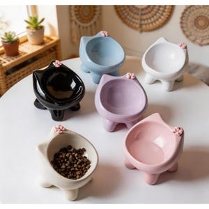Może przedstawiać: Kolekcja ceramicznych misek dla zwierząt w kształcie kota w różnych pastelowych kolorach: czarnym, niebieskim, białym, lawendowym i różowym. Każda miska ma małą różową ozdobę w kształcie kwiatka i jest wypełniona karmą lub pusta.