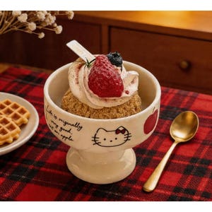 May include: A dessert bowl with a Hello Kitty design, filled with a layered dessert topped with whipped cream, a strawberry, and blueberries. A gold spoon rests beside the bowl on a red plaid surface. The bowl has the text "Life is originally very good, eat an apple."