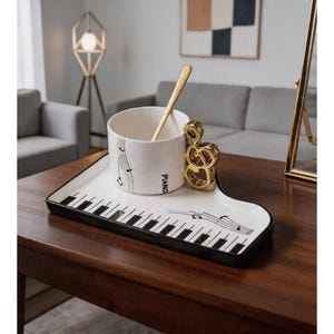 Może przedstawiać: Biały ceramiczny zestaw filiżanki i tacy z motywem klawiszy fortepianu. Na filiżance znajdują się nuty i napis "PIANO". Złota łyżeczka spoczywa w filiżance. Złoty uchwyt w kształcie klucza wiolinowego jest przymocowany do tacy.