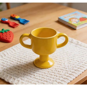 Może przedstawiać: Jasnożółty ceramiczny kubek w kształcie trofeum z dwoma uchwytami, ustawiony na białej dzianinowej podkładce. Inne przedmioty to czerwony przedmiot w kształcie truskawki, niebieski i czerwony widelec oraz książka dla dzieci.