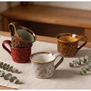 May include: A selection of ceramic mugs with a rustic aesthetic. The mugs feature speckled finishes in shades of red, brown, and grey. Each mug has a curved handle and a unique glaze. The mugs are displayed on a neutral surface.