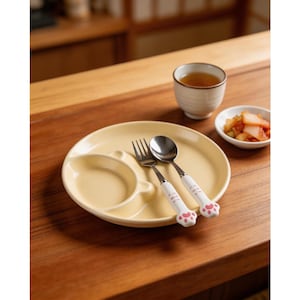 May include: A yellow divided plate with a fork and spoon set. The utensils have white handles with pink paw prints. A small bowl of kimchi and a cup of tea are also on the wooden table.