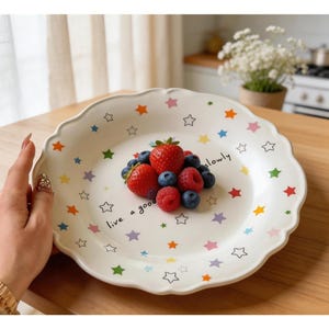 Può includere: Piatto in ceramica bianca con bordo smerlato, decorato con stelle colorate e le parole "live a good slowly". Il piatto contiene fragole, mirtilli e lamponi. Una mano con un anello d'oro tiene il piatto.
