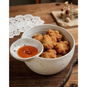 Könnte beinhalten: Eine weiße Keramikschale gefüllt mit goldbraunen, frittierten Speisen, begleitet von einer kleinen, angebrachten Schale mit oranger Soße. Die Schale steht auf einer Holzoberfläche, was auf eine Essumgebung hindeutet.