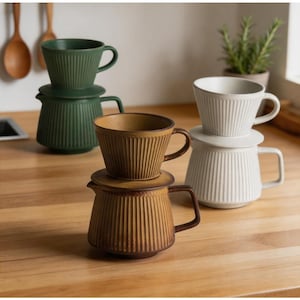 Può includere: Una collezione di set per versare il caffè in ceramica in vari colori, tra cui verde, marrone e bianco. Ogni set presenta un design a coste sulla tazza e sulla caraffa, con un coperchio abbinato. I set sono disposti su una superficie di legno.