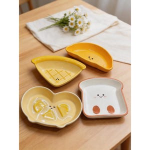 Puede incluir: Un conjunto de cuatro platos de cerámica con forma de alimentos. Los platos son amarillos, blancos y naranjas, con caras y diseños de dibujos animados. Las formas incluyen una porción de pizza, una rebanada de pan tostado, un limón y una media luna.