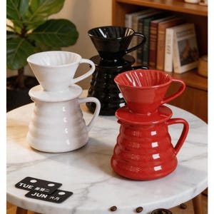 Puede incluir: Colección de cafeteras de cerámica para verter en blanco, negro y rojo. Cada juego incluye un gotero y una jarra con asa. Las cafeteras están dispuestas sobre una superficie similar al mármol, con un calendario que muestra "TUE" y "JUN".