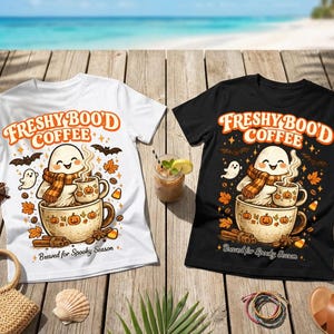 以下が含まれることがあります： 白いTシャツと黒いTシャツ。どちらも、大きなマグカップの中でコーヒーマグを持つゴーストのグラフィックが描かれています。「FRESHY BOO'D COFFEE」と「Brewed for Spooky Season」という文字が、秋の葉、コウモリ、シナモンスティックのイラストと共にデザインされています。
