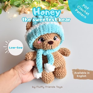 May include: A brown plush crochet bear toy wearing a light blue and white striped beanie and a matching light blue scarf with white pom-poms. The bear has large dark eyes and pink cheeks. Text on the image reads Honey the sweetest bear, Low-Sew, PDF Crochet Pattern, Available in English, and by Fluffy Friends Toys.
