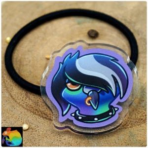 Könnte beinhalten: Ein schwarzes elastisches Haargummi mit einem durchsichtigen Acryl-Anhänger, der eine Zeichentrickfigur mit blauer Haut, dunklem Haar mit weißer Strähne, einem orangefarbenen Auge, einem Nasenring und einem schwarzen Stachelhalsband zeigt.