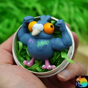 Könnte beinhalten: Eine kleine, blaue, muskulöse Taubenfigur mit großen orangefarbenen Augen, einem weißen Schnabel und rosa Füßen. Der Vogel ist mit angewinkelten Armen dargestellt und hat grüne und violette Markierungen auf der Brust, in einem runden Behälter.