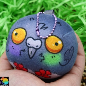 Puede incluir: Un peluche redondo azul con forma de paloma, con grandes ojos amarillos y naranjas, un pico blanco y detalles de flores rojas. Tiene una cara morada y verde y una pequeña cadena morada para colgar.