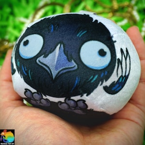 Può includere: Un peluche rotondo con un'illustrazione stampata di un uccello nero con grandi occhi spalancati, un becco appuntito e piccoli piedi. L'uccello è su uno sfondo bianco e il testo Rainbow Rat Art è stampato nell'angolo in basso a sinistra.