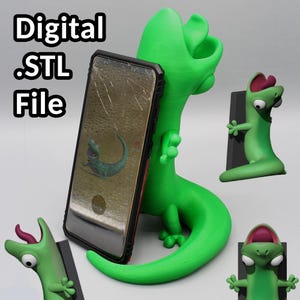 May include: A bright green 3D-printed lizard phone stand holding a smartphone with a cracked screen. The phone displays a cartoon lizard. The words "Digital .STL File" are in the upper left corner. Additional green lizard figurines are in the background.