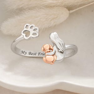 Puede incluir: Anillo plateado con diseño de huella de pata y la inscripción "My Best Friend". El anillo presenta un perro color oro rosa abrazando una figura plateada. El anillo está sobre una superficie blanca.