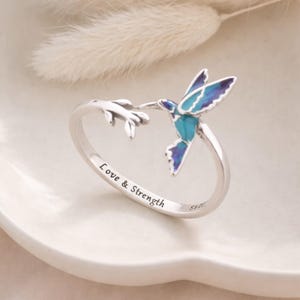 May include: A silver ring featuring a hummingbird design with blue and teal enamel detailing. The ring has the words "Love & Strength" engraved on the band. The hummingbird is perched on a branch.