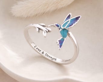 Könnte beinhalten: Silberner Ring mit einem Kolibri-Design, mit blau-türkisfarbenem Emaille. Der Ring trägt die Inschrift "Love & Strength". Der Kolibri sitzt auf einem Ast. Ein filigranes Schmuckstück.