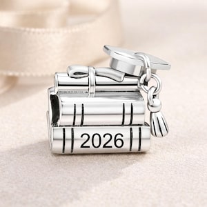 Pode incluir: Um charm de prata com uma pilha de livros com o ano "2026" gravado na lateral. Um chapéu de formatura está em cima dos livros, com um borla pendurada. O charm foi projetado como um presente de formatura.