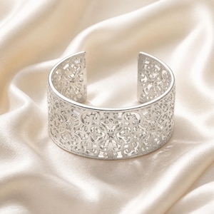 May include: A silver-toned cuff bracelet featuring an intricate, openwork filigree pattern throughout the entire band. The metal has a polished, reflective finish, and the design consists of repeating geometric shapes and scrolling motifs.