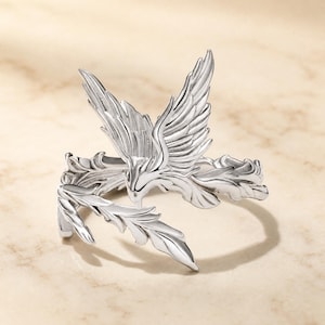 May include: A silver-toned ring featuring a detailed bird with outstretched wings and a long, curved tail that wraps around the finger. The metal surface has a polished finish with engraved lines defining the feathers and wings of the bird.