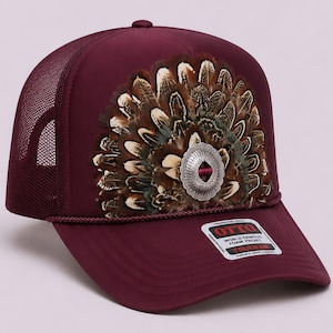May include: A wine-colored trucker hat featuring a fan-shaped arrangement of brown, black, and white feathers centered with a silver-toned metal concho. The cap includes a mesh back, a cord trim above the brim, and an Otto brand sticker on the visor.