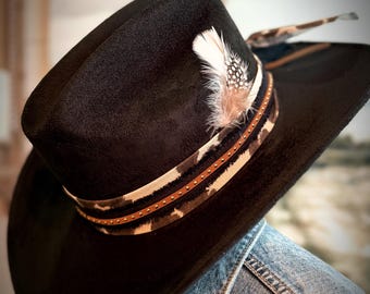 May include: Black cowboy hat with a decorative band featuring a brown and white pattern and small studs. A feather is attached to the side of the hat, adding a rustic touch. The hat is worn over a denim jacket.