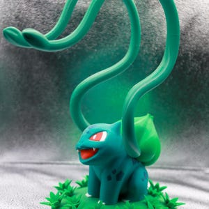 May include: A teal and green Bulbasaur figurine with red eyes and a smiling mouth. The figurine has a large green bulb on its back and is surrounded by green leaves. The figure has long green vines extending upwards.