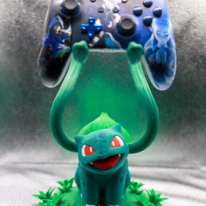 May include: A teal and green Bulbasaur figure holding up a video game controller. The controller is black with blue accents and features character art. The Bulbasaur is on a green base with leaf details.