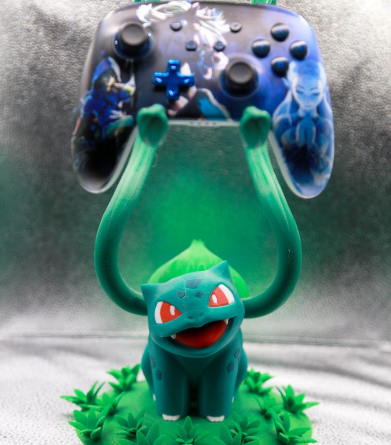 May include: A teal and green Bulbasaur figure holding up a video game controller. The controller is black with blue accents and features character art. The Bulbasaur is on a green base with leaf details.