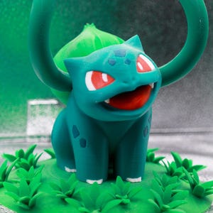 May include: A teal Bulbasaur figurine with red eyes and an open mouth, set on a green base with grass details. A green vine-like structure arches above the figure. The background is a gradient of green and silver.