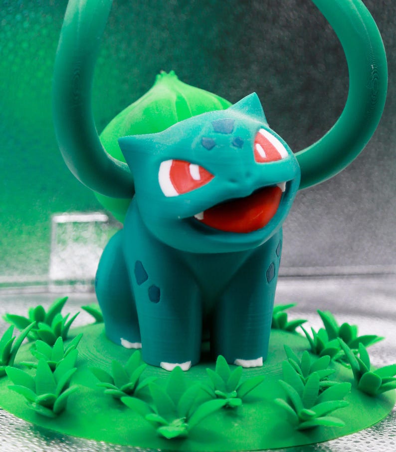 May include: A teal Bulbasaur figurine with red eyes and an open mouth, set on a green base with grass details. A green vine-like structure arches above the figure. The background is a gradient of green and silver.