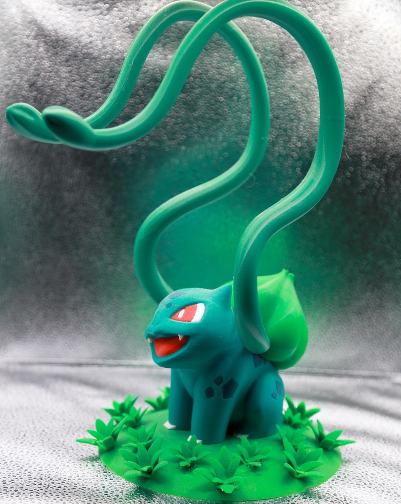 May include: A teal and green Bulbasaur figurine with red eyes and a smiling mouth. The figurine has a large green bulb on its back and is surrounded by green leaves. The figure has long green vines extending upwards.
