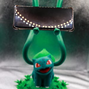 May include: A green Bulbasaur figure holding a black rectangular object with a row of small, evenly spaced, yellow lights. The Bulbasaur has red eyes and is sitting on a bed of green foliage.