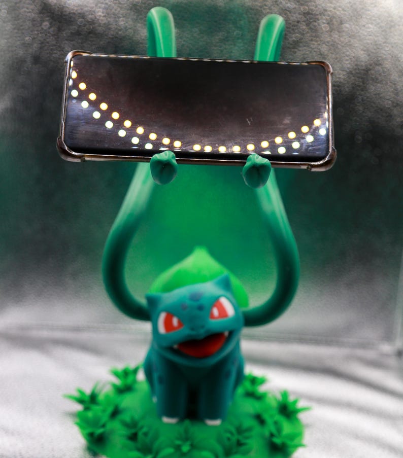 May include: A green Bulbasaur figure holding a black rectangular object with a row of small, evenly spaced, yellow lights. The Bulbasaur has red eyes and is sitting on a bed of green foliage.