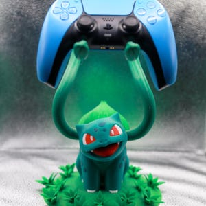 May include: A teal Bulbasaur figure holding up a blue and black PlayStation 5 controller. The Bulbasaur is on a green base with grass details. The controller has white button symbols.