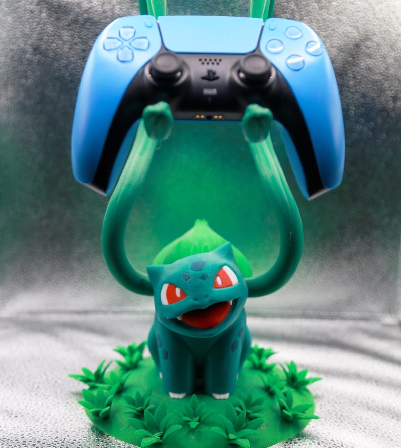 May include: A teal Bulbasaur figure holding up a blue and black PlayStation 5 controller. The Bulbasaur is on a green base with grass details. The controller has white button symbols.
