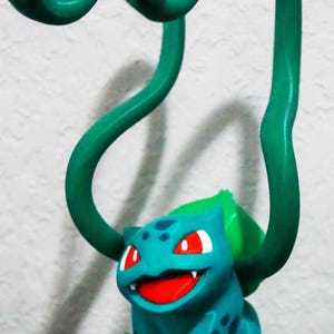 May include: A teal Bulbasaur figurine with red eyes and a smiling mouth, set on a green base with leafy accents. Two green vines arch over the figure. A fun, decorative item.