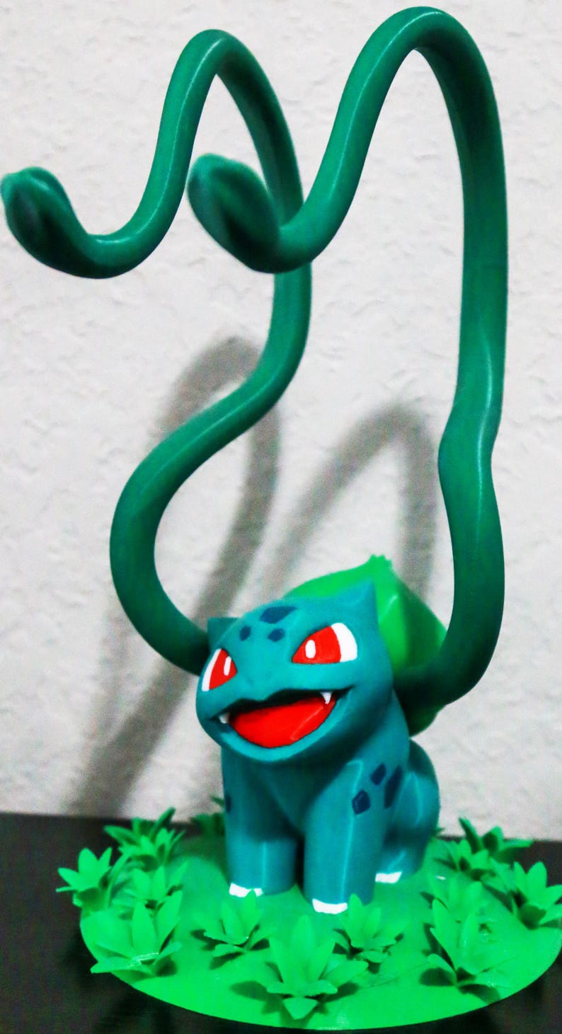 May include: A teal Bulbasaur figurine with red eyes and a smiling mouth, set on a green base with leafy accents. Two green vines arch over the figure. A fun, decorative item.