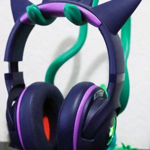 May include: Dark purple gaming headset with purple and green accents, resting on a green stand. The headset has ear-shaped design elements and a built-in microphone. The ear cups are dark purple with a red interior.