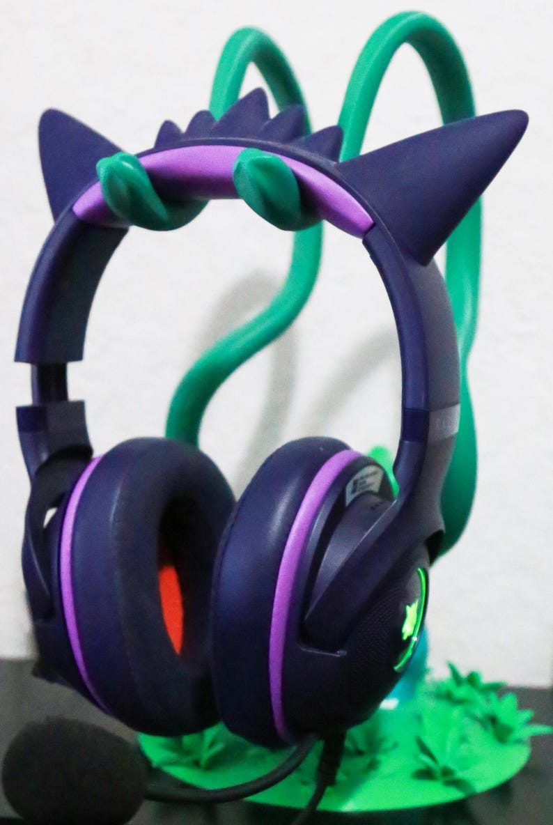 May include: Dark purple gaming headset with purple and green accents, resting on a green stand. The headset has ear-shaped design elements and a built-in microphone. The ear cups are dark purple with a red interior.