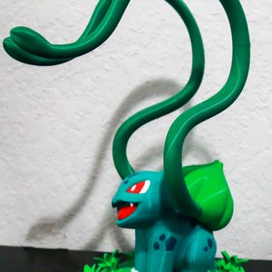 May include: A 3D-printed figurine of a teal creature with red eyes and a red mouth, featuring green vines and a green leaf on its back. The figurine is set on a green base with leaf-shaped details.