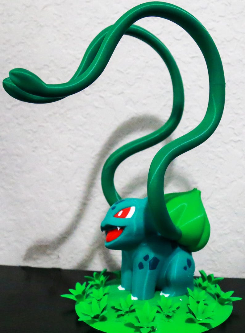 May include: A 3D-printed figurine of a teal creature with red eyes and a red mouth, featuring green vines and a green leaf on its back. The figurine is set on a green base with leaf-shaped details.